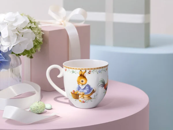 Una taza decorativa Annual Easter Edition 2026 con diseño de conejo sobre una mesa rosa junto a una caja de regalo y un jarrón con flores blancas.