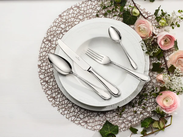 Mademoiselle cutlery with pastel-coloured roses A set of Villeroy & Boch Mademoiselle white plates with a knife, a fork and two spoons elegantly arranged on a lace doily, with pink and white flowers adding a decorative touch to the table setting.