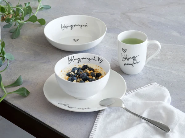 Cuenco Statement "Lieblingsmensch" lleno de arándanos El cuenco Statement "Lieblingsmensch" con arándanos, plato, taza y cuchara a juego sobre una mesa.