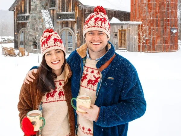 McCallister Sweaters A smiling couple in matching winter hats and sweaters stands outside in the snow, holding mugs, with a rustic barn and silo in the background.