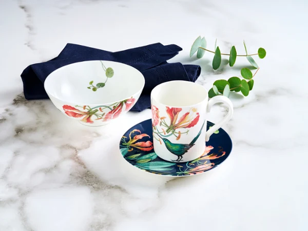 aus-avarua-indoor-nofood-nopeople-giftset7085 Avarua set for one, featuring a cup, saucer, and small bowl, is beautifully adorned with delicate fauna and red floral accents, and complemented by a rich dark blue napkin.