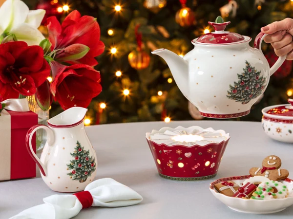 A festive table with a teapot, a warmer, a cup, Christmas ceramics, cookies, and a decorated tree in the background.