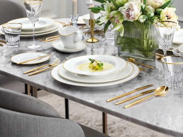 Elegant table set on a marble surface with gold-rimmed plates, gold flatware, crystal glasses, a vase of white flowers, and a soup served on the central plate.