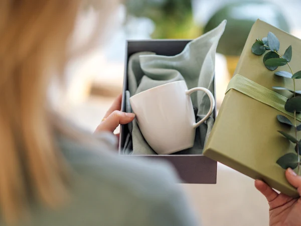 Taza Crafted de cerámica blanca en caja de regalo Mujer sosteniendo una caja de regalo con una taza de cerámica Crafted y un paño verde decorado con una rama de eucalipto.