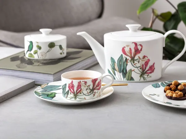 Avarua Tee- Set auf einem Tisch Eine Teekanne, eine Tasse und ein Teller mit Snacks im Blumenmuster sind auf einem Tisch neben Büchern und einer Pflanze im Hintergrund angeordnet.