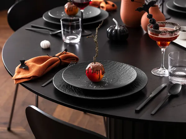 Une table de salle à manger noire est recouverte d’assiettes noires Manufacture, de serviettes orange ainsi que de boissons et de décorations sur le thème d’Halloween, dont une pomme enrobée de chocolat.