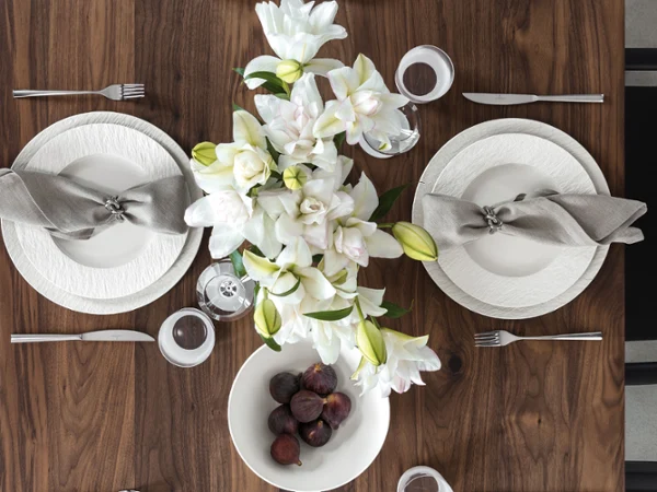 Manufacture Rock Blanc festive table setting with white plates Overhead flat lay of a dark walnut wood dining table elegantly set with textured white Manufacture Rock Blanc porcelain plates, grey linen napkins with silver rings, a natural centerpiece of white lilies, and a white bowl filled with fresh, dark figs.