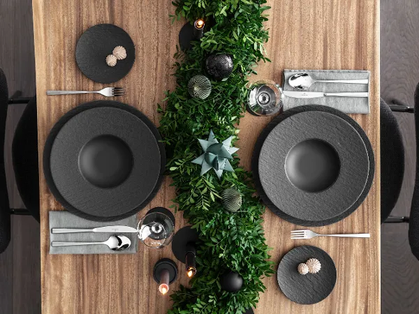 Black Manufacture plates on a modern Christmas table A beautiful dining table setting with black Manufacture plates, grey napkins and silver cutlery. Decorative green foliage runs along the table, accentuated by candles and decorative items.