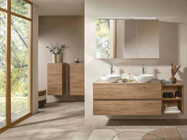 Arto wood-look washstand and a white washbasin Modern bathroom with two countertop washbasins on a wooden washstand, wall cabinets, round mirrors and a view of trees through a large window.