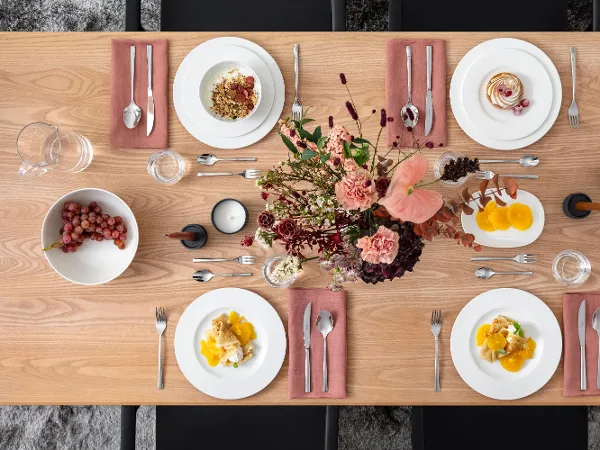 Une table rectangulaire en bois est dressée pour quatre personnes. Il y a des assiettes avec de la nourriture, des serviettes roses, des couverts, des bougies et une composition florale. Des raisins et des épices sont également présents.