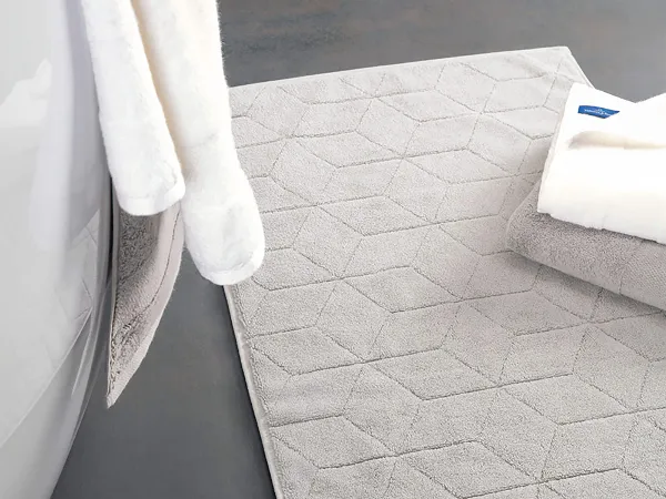 Badematte Carré in French Linen neben einer weißen Badewanne Graue Badematte mit geometrischem Muster auf dunklem Boden neben einer weißen Badewanne und weißen Handtüchern. Grüne Pflanze im Vordergrund.