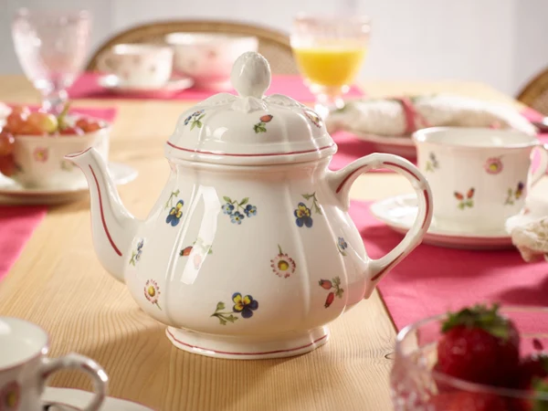 A floral-patterned teapot is centered on a wooden table set with pink placemats, cups, and fruit, creating a warm and inviting breakfast scene.