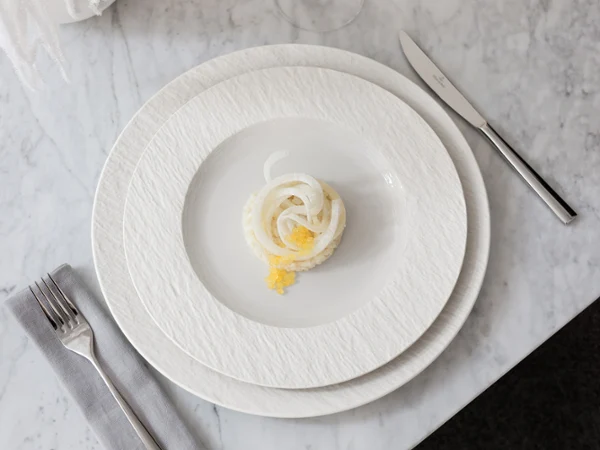 A small portion of food is arranged on a white Manufacture plate with a knife, fork, and napkin on a marble surface.