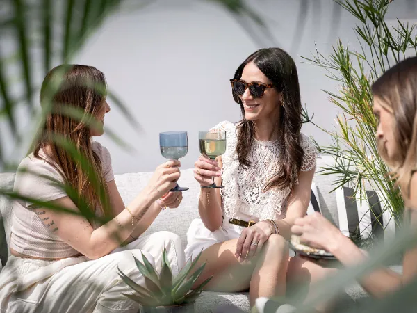 Women toasting with Like glasses Three women sitting outside on a beige sofa, smiling and holding up Like glasses of wine. They are surrounded by green plants and appear to be enjoying a sunny day.