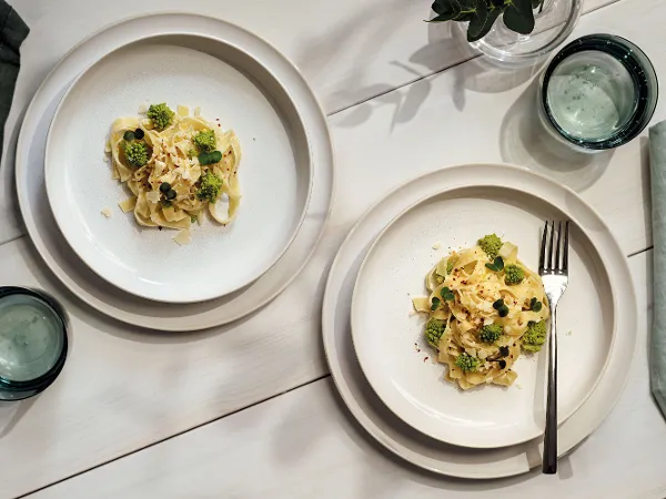 Piatto Crafted con pasta e pianta in vaso su una tavola bianca Due piatti Crafted con broccoli e salsa su una tavola bianca, con bicchieri d'acqua e una piccola pianta in vaso.