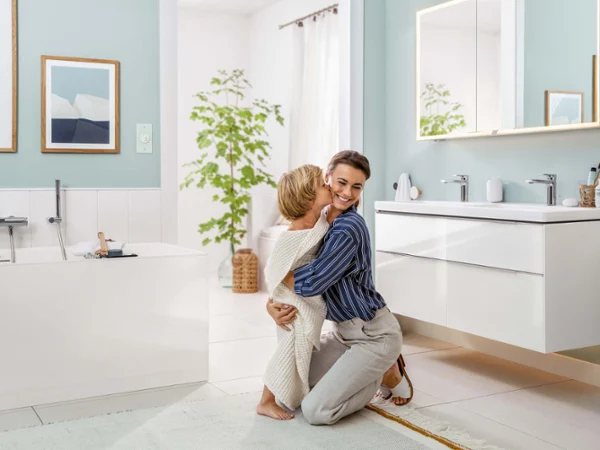 Łazienka rodzinna Subway 3.0 firmy Villeroy & Boch Kobieta klęczy i uśmiecha się, przytulając dziecko, w jasnej, nowoczesnej łazience. W tle widać wannę, umywalkę i zieloną roślinę.