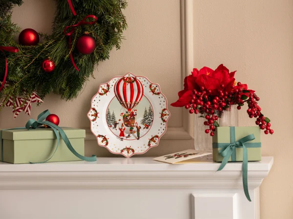 Annual Chistmas edition 2026 plate A Christmas wreath, a decorative plate featuring a hot-air balloon by Villeroy & Boch, green gift boxes, and red flowers, arranged on a white mantelpiece.