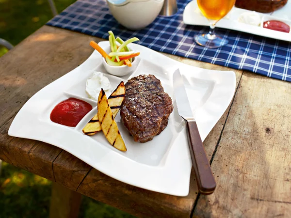Płyta grillowa NewWave ze stekiem i ziemniakami Grillowany stek leży na białym talerzu do grillowania NewWave z grillowanymi plastrami ziemniaków, porcją kwaśnej śmietany, ketchupem i pokrojonymi warzywami. Na krawędzi talerza spoczywa teksański nóż do steków. W tle znajduje się napój.