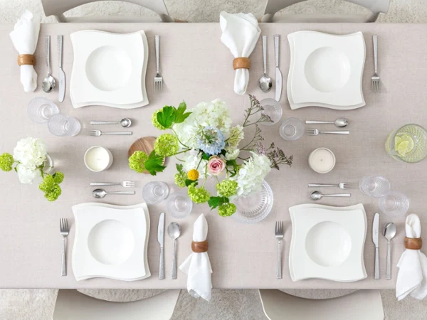 NewWave festively decorated A rectangular dining table for four people with white square plates, silver cutlery, glasses, cream-coloured napkins with wooden rings, a floral arrangement, candles, and a jug of lemon water.