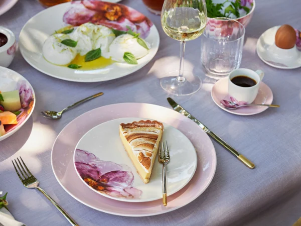Una tavola apparecchiata per il brunch con le stoviglie di Rose Garden Ambiente elegante per il brunch con stoviglie della serie Rose Garden, una fetta di torta su un piatto Rose Garden, macedonia di frutta fresca, caffè e un bicchiere di vino bianco.