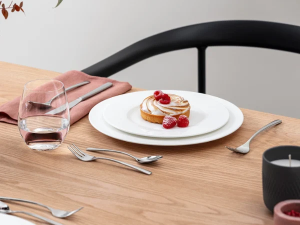 Royal combined with sweet delicacies A dessert garnished with raspberries stands on a royal plate with cutlery on a wooden table. Next to it is a glass of water, a pink napkin and a flower arrangement.