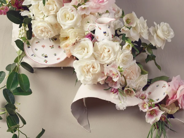 Elegant floral arrangement featuring white and pink roses intertwined with green leaves, surrounded by a floral-patterned plate and pitcher. The scene conveys a romantic, vintage vibe.