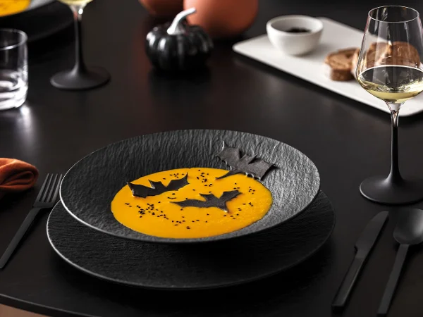 Un piatto nero con zuppa arancione e guarnizioni a forma di pipistrello su una tavola scura con un bicchiere da vino, posate nere e un piatto bianco con cibo sullo sfondo.