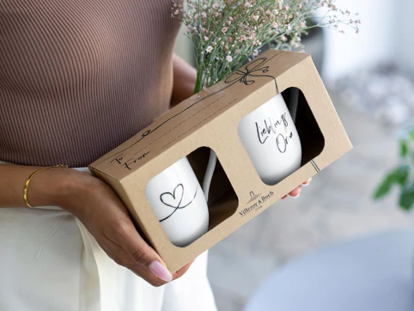 Two Statement mugs with the message "Lieblingsoma" in a gift box A person holding a box with two Statement mugs with the message "Lieblingsoma" and a bouquet of small flowers.