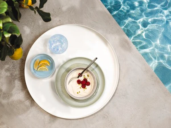 A round tray with tableware from the Perlemor Aqua and Perlemor Alga collection A round tray with tableware from the Perlemor Aqua collection and a glass of water, at the edge of a pool.