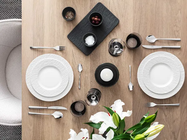 Elegant dining table with white Manufacture plates, Piemont cutlery and decorative flowers on a wooden surface.