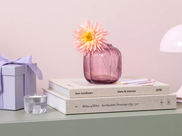 Een roze Fleur-vaas met een enkele bloem staat op gestapelde boeken naast een geschenkdoos en een kleine lamp op een groene plank.