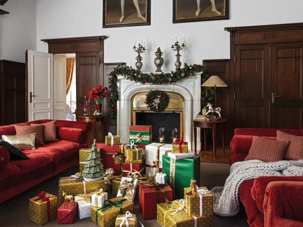 Christmas presents piled up in a living room A cosy living room with red sofas, a fireplace decorated with garlands and candles from Villeroy & Boch and lots of wrapped Toy’s Christmas presents on the floor.