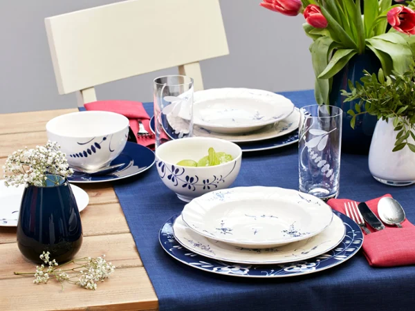 A dining table with Old Luxembourg crockery on a blue table runner next to a Drop vase with red tulips and green plants.