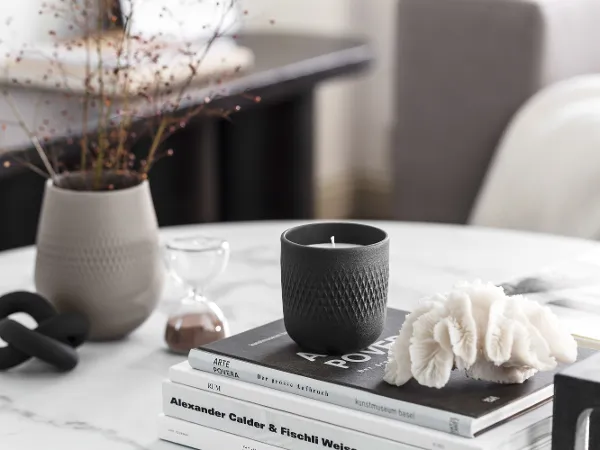 A Manufacture scented candle and vase on a marble table Elegant home decor arrangement with a black Manufacture scented candle, books and a light-grey Manufacture vase on a marble table.