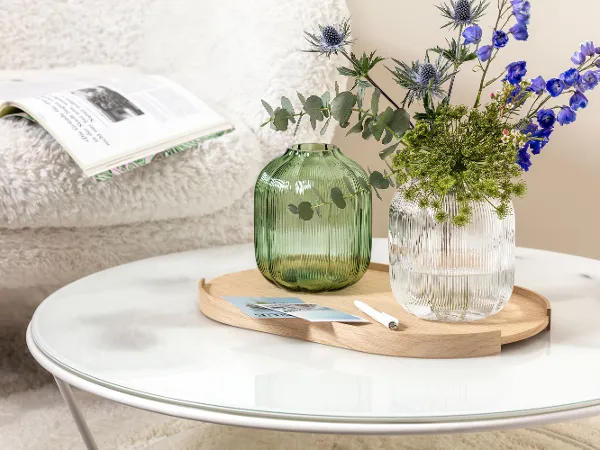 Ein runder Couchtisch mit einer Villeroy & Boch Fleur Vase, die blaue und grüne Blumen hält. Ein runder Couchtisch mit einer Villeroy & Boch Fleur Vase, die blaue und grüne Blumen hält. Im Hintergrund liegt eine aufgeschlagene Zeitschrift auf einem weißen, flauschigen Stuhl.