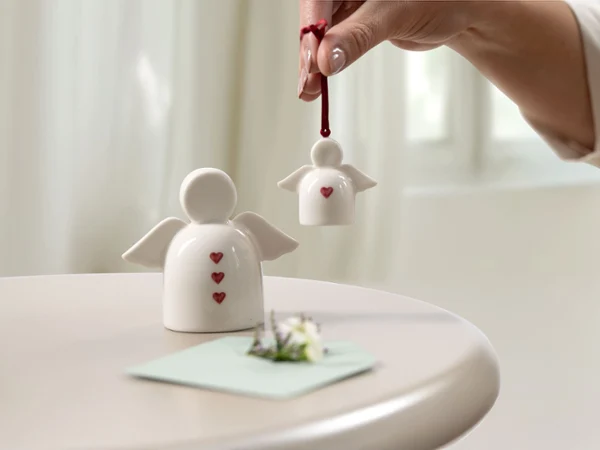 Ozdoby anielskie z czerwonymi motywami serca od with love jako pomysły na prezent Ręka trzyma małą ceramiczną ozdobę anielską z czerwonym motywem serca od with love obok większej figurki anioła od with love i kartki na stole, co stanowi inspirację na prezent.