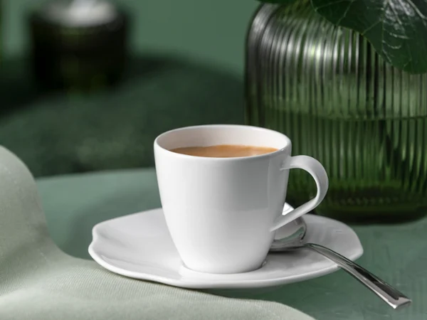 Eine weiße Pura Kaffeetasse mit Untertasse und Löffel steht neben einer grünen Glasvase Eine weiße Pura Kaffeetasse mit Untertasse und Löffel steht auf einer grünen Fläche neben einer grünen Glasvase.