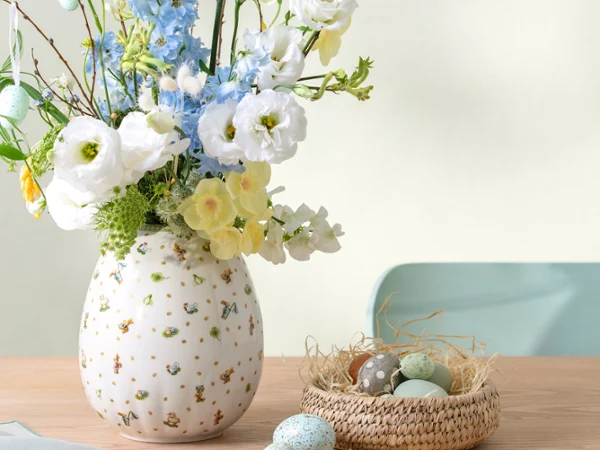 A Easter Delight vase with white and blue flowers sits beside a small nest filled with pastel eggs on a wooden table.