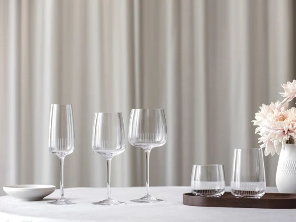 Elegant table setting with four crystal glasses and a white vase of pink flowers on a white tablecloth against a soft, neutral background.