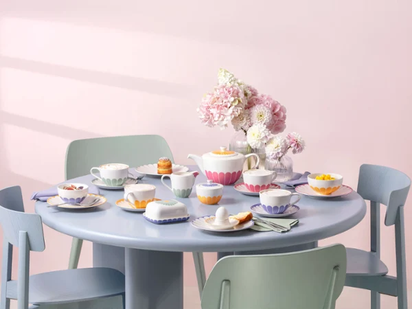 Runder pastellblauer Tisch mit pastellfarbenen Stühlen, gedeckt für Tee, mit Tassen und Teller von Fleur mit floralen Mustern, feines Gebäck und eine Vase mit rosa Blumen als Mittelpunkt.