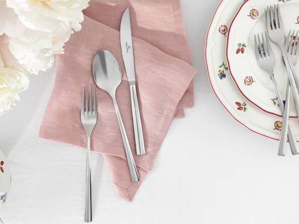 Piemont cutlery lies on a pink Lifestyle Accessories napkin next to stacked Petite Fleur plates on a white tablecloth.