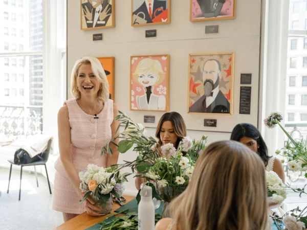 A woman, Dorinda Medley, in a pink dress smiles while arranging flowers on a table with three others. Colorful portraits hang on a wall, with large windows in the background.