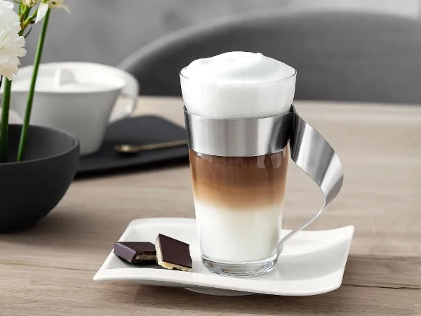 Un latte stratifié dans un verre NewWave avec une anse en métal est posé à côté de deux morceaux de chocolat sur une assiette blanche de Villeroy & Boch NewWave. Au fond, sur la table en bois, il y a des fleurs blanches et une tasse.