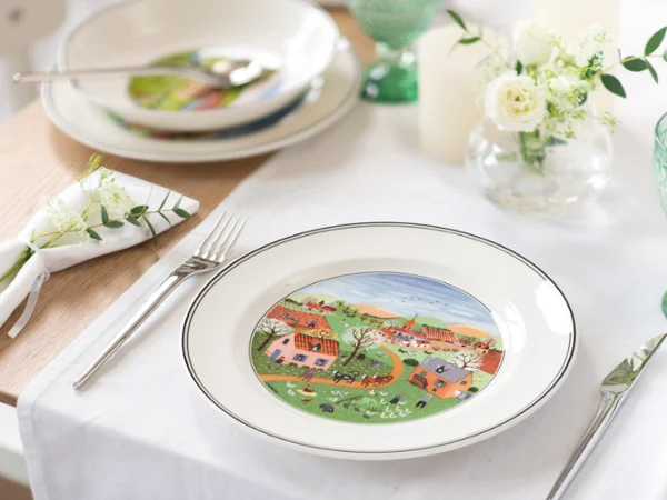 Elegant table set with white dishes, featuring a colorful countryside scene with houses and trees. Accented by green glassware and white floral decor.
