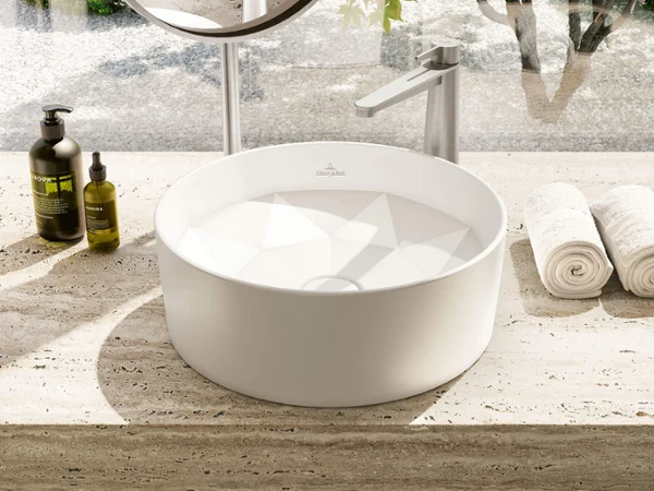 Round white sink on a speckled countertop with rolled towels, soap bottles, and a round mirror above.