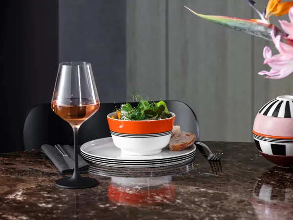 Memphis von Villeroy & Boch ideal für Vorspeisen Ein Esszimmer mit einer Memphis Salatschüssel, einem Weinglas, gestreiften Memphis Tellern, einer tropischen Blume in einer Memphis Vase und einer Tischplatte aus Marmor.