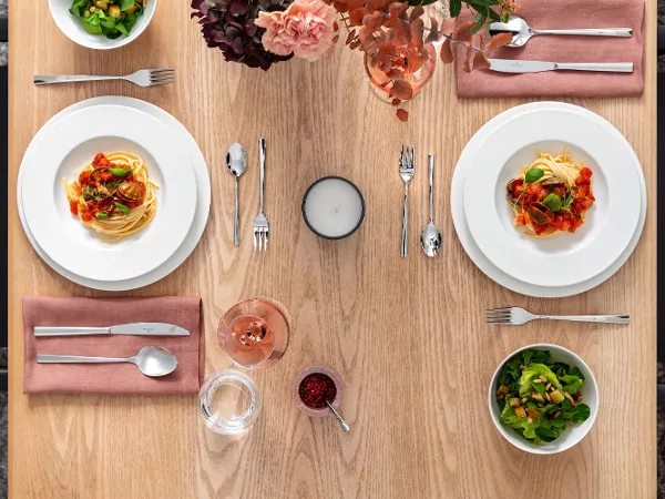 Table de salle à manger avec deux assiettes royales de pâtes, deux salades, des couverts, une bougie et une boisson. Les assiettes Villeroy & Boch sont posées sur des serviettes roses et une composition florale rose est disposée sur la table.	