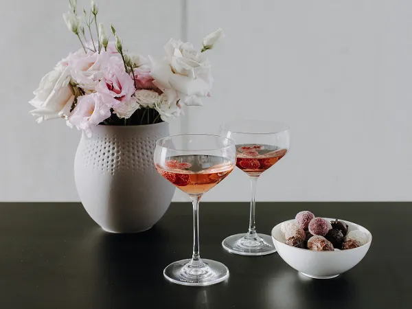 Purismo Gläser, Manufacture Schale und Manufacture Vase Zwei Gläser Roséwein, eine weiße Vase mit Blumen und eine Schale Pralinen auf einem dunklen Tisch.