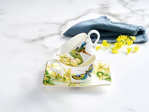 Amazonia tea set for two, arranged on a rectangular saucer adorned with vibrant fauna and delicate yellow floral accents, complemented by a blue linen napkin.