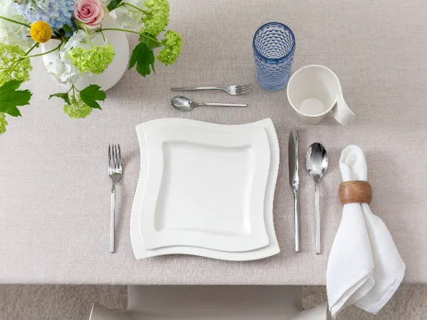 Clean, modern top view of a single table setting featuring NewWave porcelain plates and cup, Boston Blue long drink glass, and matching NewWave cutlery. Neutral tones and minimalist aesthetic create a contemporary dining impression.